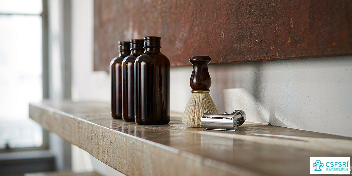 Three dark-amber apothecary-style skincare bottles on a concrete shelf, with a single shaving brush and stainless steel razor in soft daylight